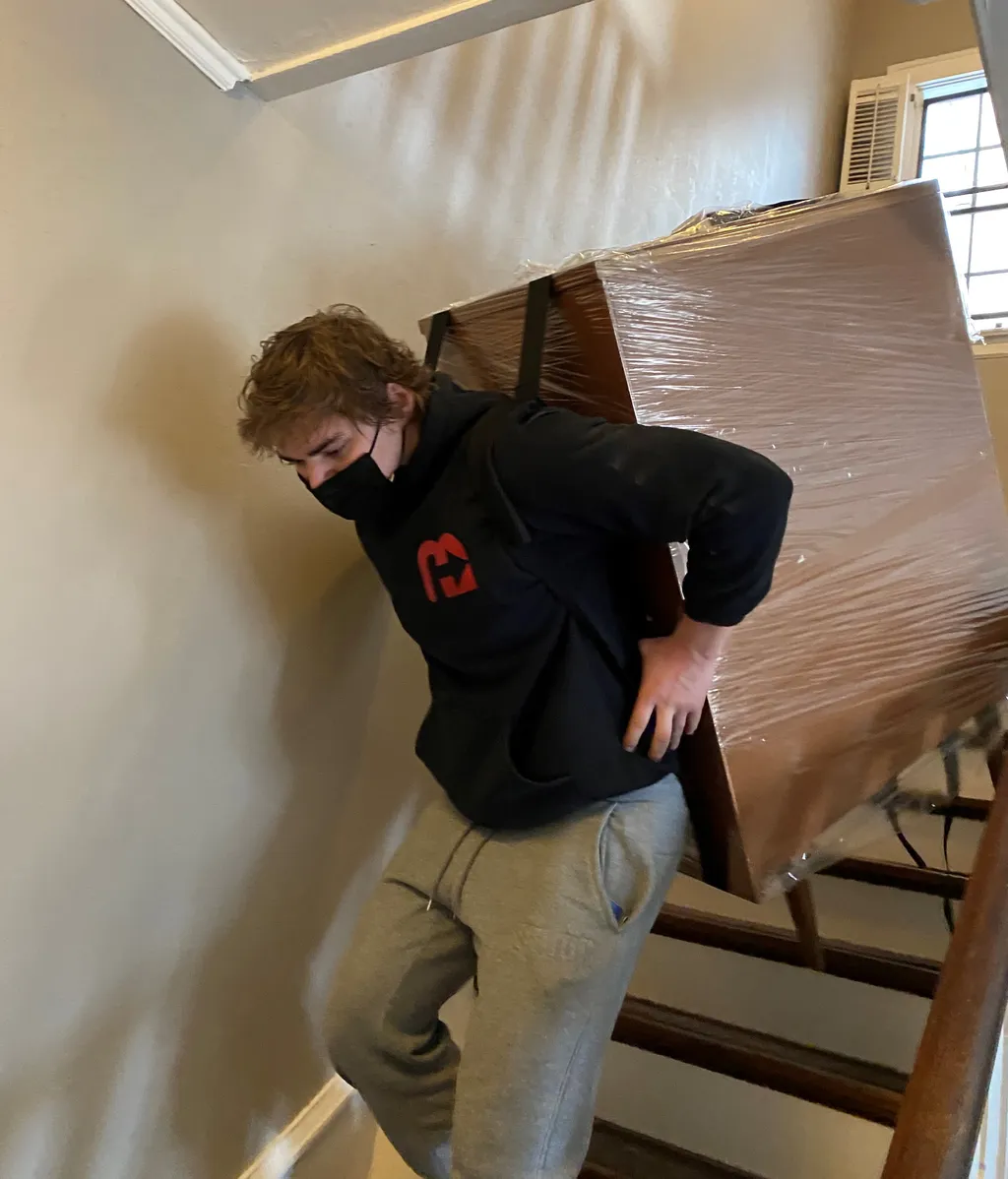 Professional mover in black uniform carrying a plastic-wrapped piece of furniture up a residential staircase