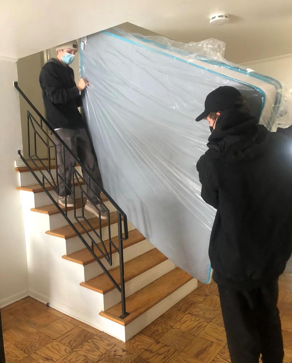 Two Monolith movers carrying a plastic-wrapped mattress down a residential staircase