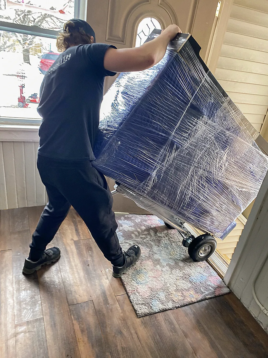 Monolith mover wheeling a plastic-wrapped appliance on a dolly through a residential doorway